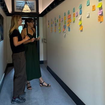 Two women in a corridor studying a wall covered with colourful sticky notes arranged in columns.